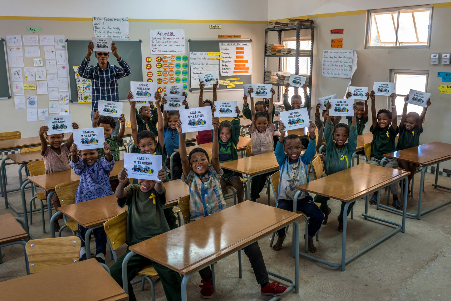 Children in a classroom holding up signs with text, with desks and educational materials around.